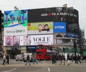 Curved bank of large digital advertising screens above a busy city junction, with a red double-decker bus and pedestrians crossing below