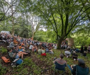 Large crowd seated on a wooded grassy slope in dappled sunlight, watching an outdoor performance