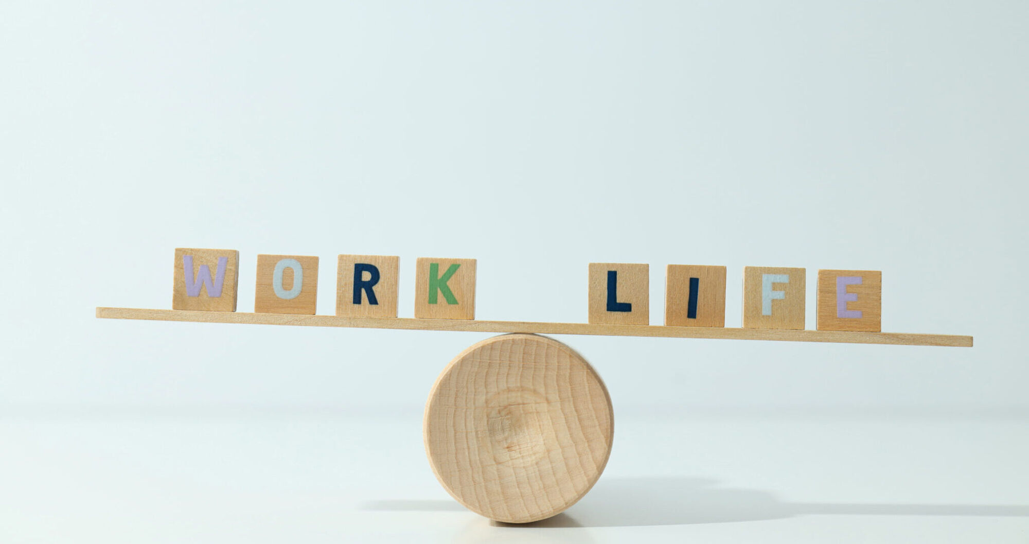 Wooden letter tiles spelling WORK and LIFE balanced on a plank resting on a round wooden block