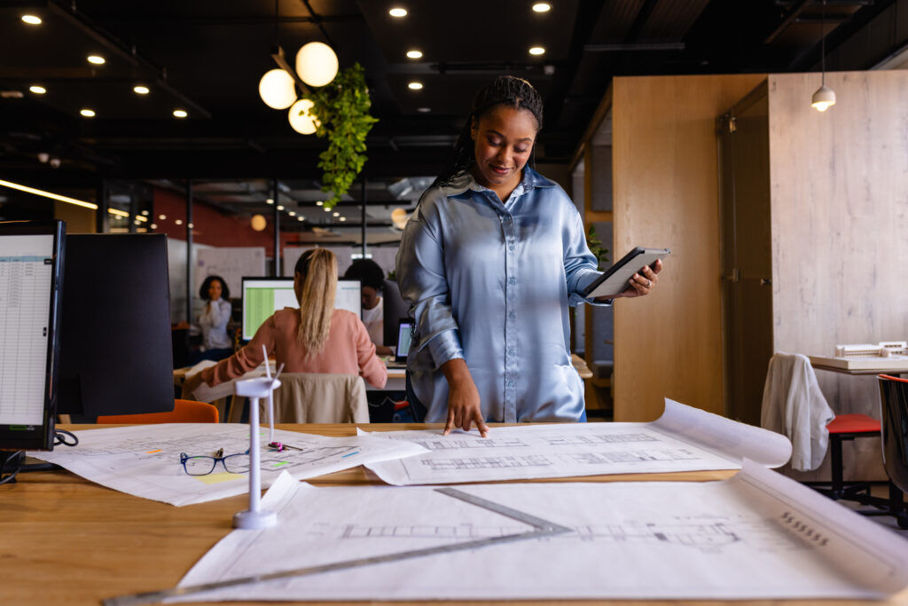 Woman holding a tablet points at large architectural floor plans spread across a desk in an open-plan office