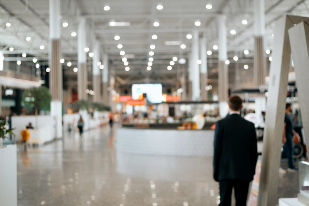 Man in a dark suit walking through a bright, softly blurred exhibition hall past white stands and a curved counter