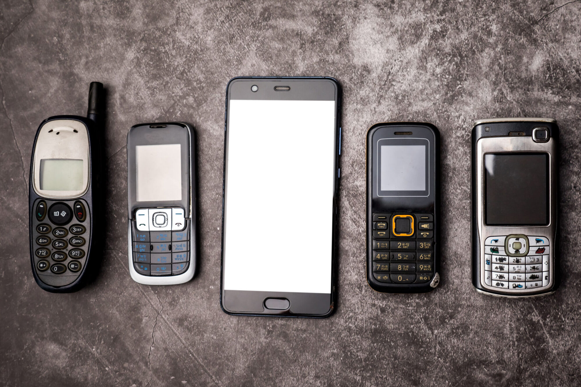 Five phones laid on grey concrete: four older keypad handsets flanking a modern blank-screen touchscreen smartphone