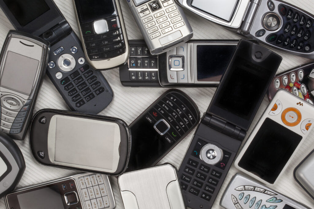 Pile of old mobile phones on a pale wooden surface, including flip phones, sliders and keypad handsets