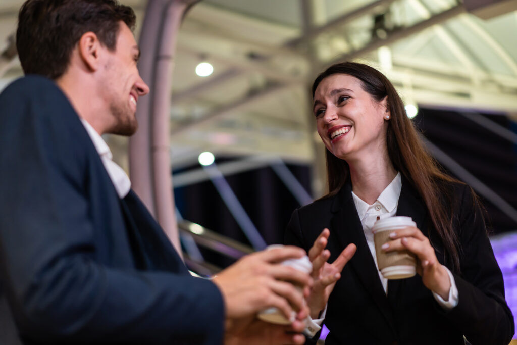 Man and woman in business suits laughing together over takeaway coffee cups at a venue