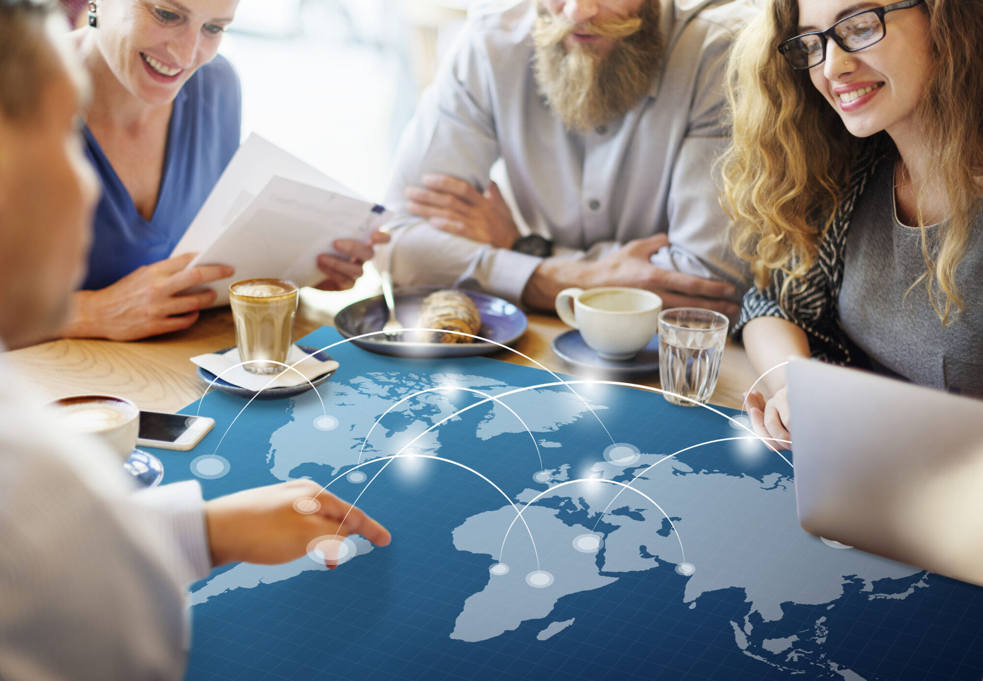 People gathered round a cafe table with coffee and papers, a world map with connecting arcs overlaid across the tabletop