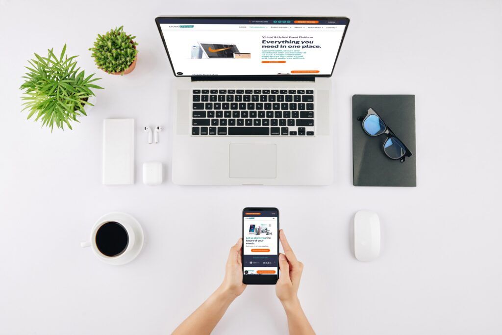 Overhead desk shot of a laptop open on the CrowdComms website while hands hold a phone showing the mobile version