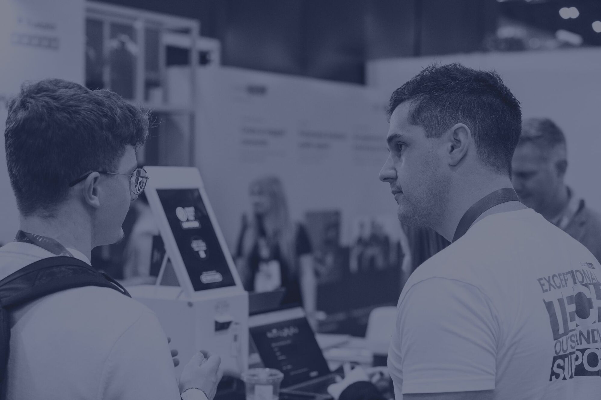 Black and white photograph of two visitors talking beside a self-service kiosk screen on an exhibition floor, with stands behind them