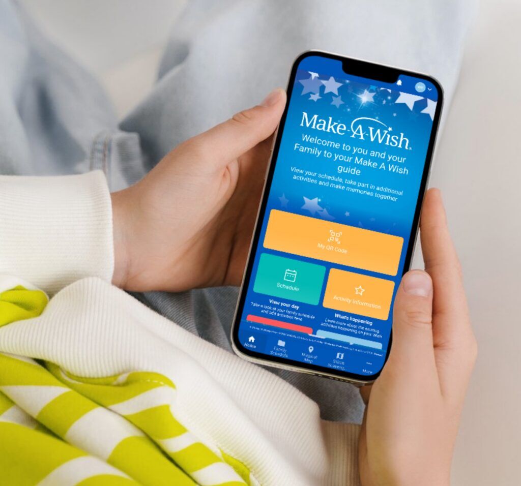 Hands holding a phone showing a Make-A-Wish app welcome screen with My QR Code, Schedule and Activity Information tiles