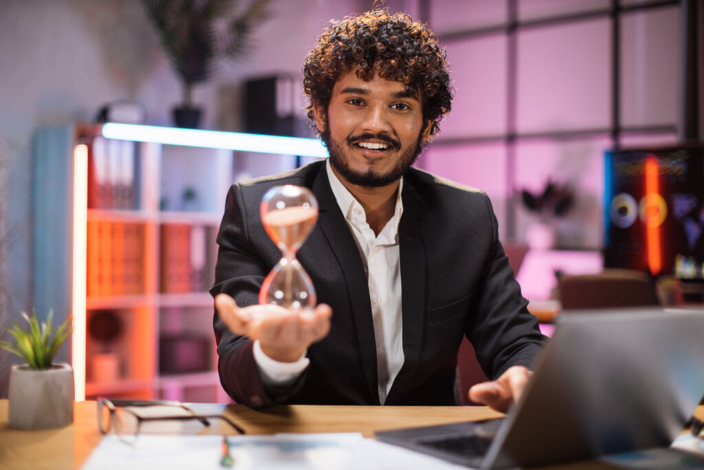 Smiling man in a suit at a desk holding out an hourglass on his open palm, in an office lit pink and orange