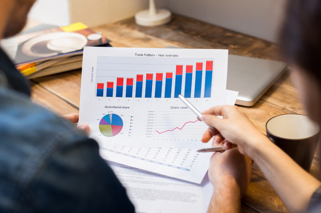 Two people at a wooden desk reviewing printed charts, including a stacked bar chart labelled "Trade Pattern - Year overview", a stock market share pie chart and a share value line graph
