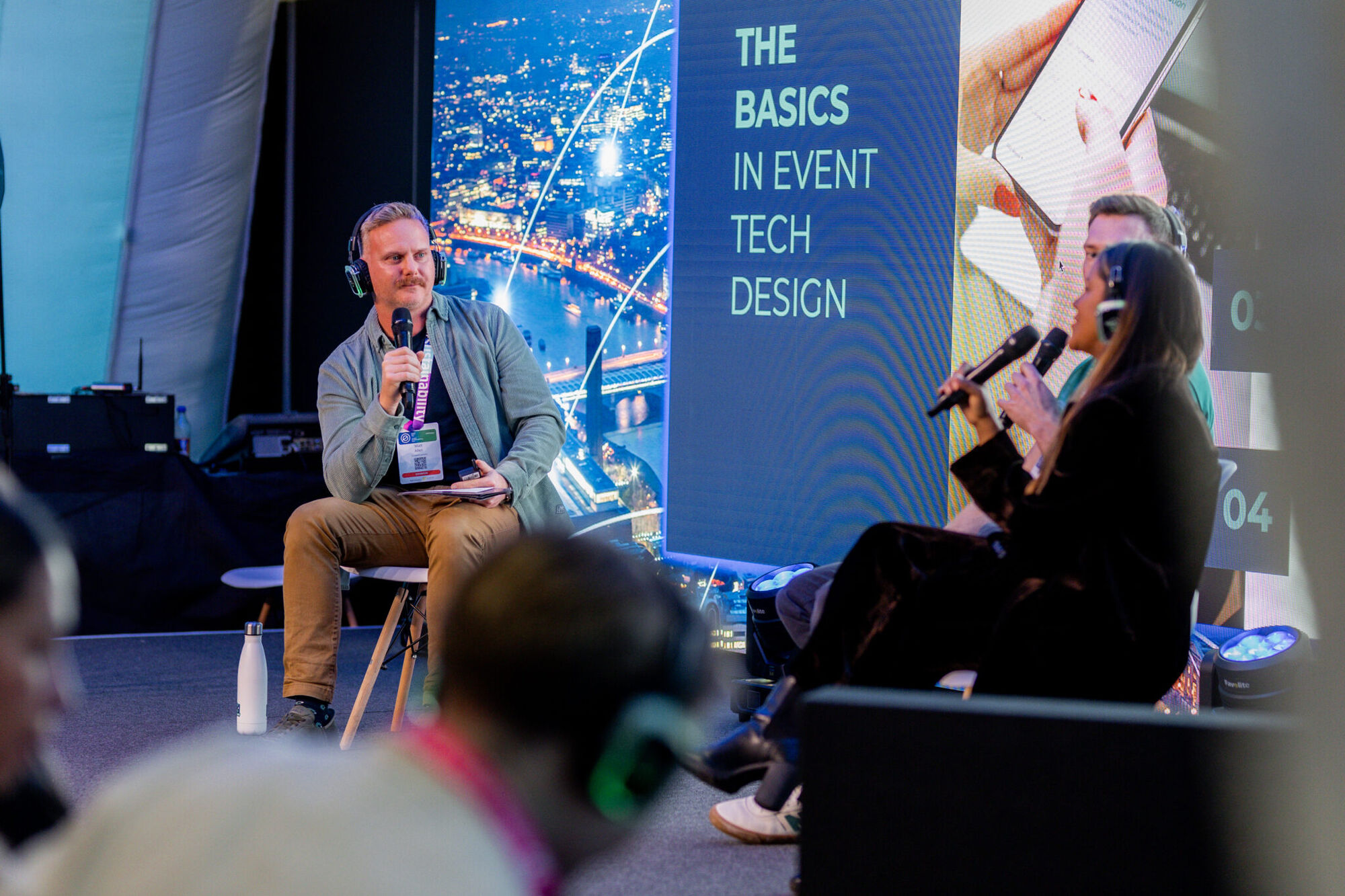 Panellist in headphones speaking into a microphone on stage, the screen behind reading "The Basics in Event Tech Design", with two panellists opposite