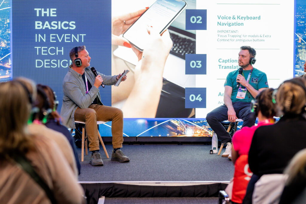 Two panellists talking on stage in front of a slide listing accessibility points including voice and keyboard navigation, and captions and translation