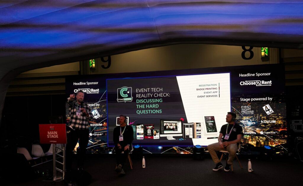 Host pointing towards the audience beside two seated panellists, the stage screen reading "Event Tech Reality Check: Discussing the Hard Questions"