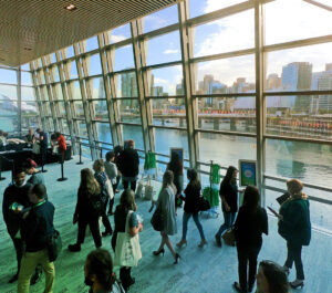 Attendees walking through a glass-walled conference concourse, with a waterfront and city skyline outside, green carpet and banner stands