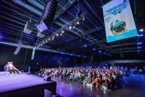 A large auditorium under blue and purple stage lighting, with a speaker on a low stage, a full seated audience and a cartoon character on the main screen