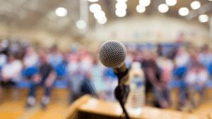 A microphone on a lectern in sharp focus, with a blurred seated audience and warm ceiling lights behind