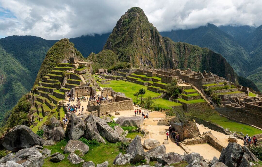 The stone terraces and ruins of Machu Picchu with visitors walking through, backed by cloud-covered green mountain peaks