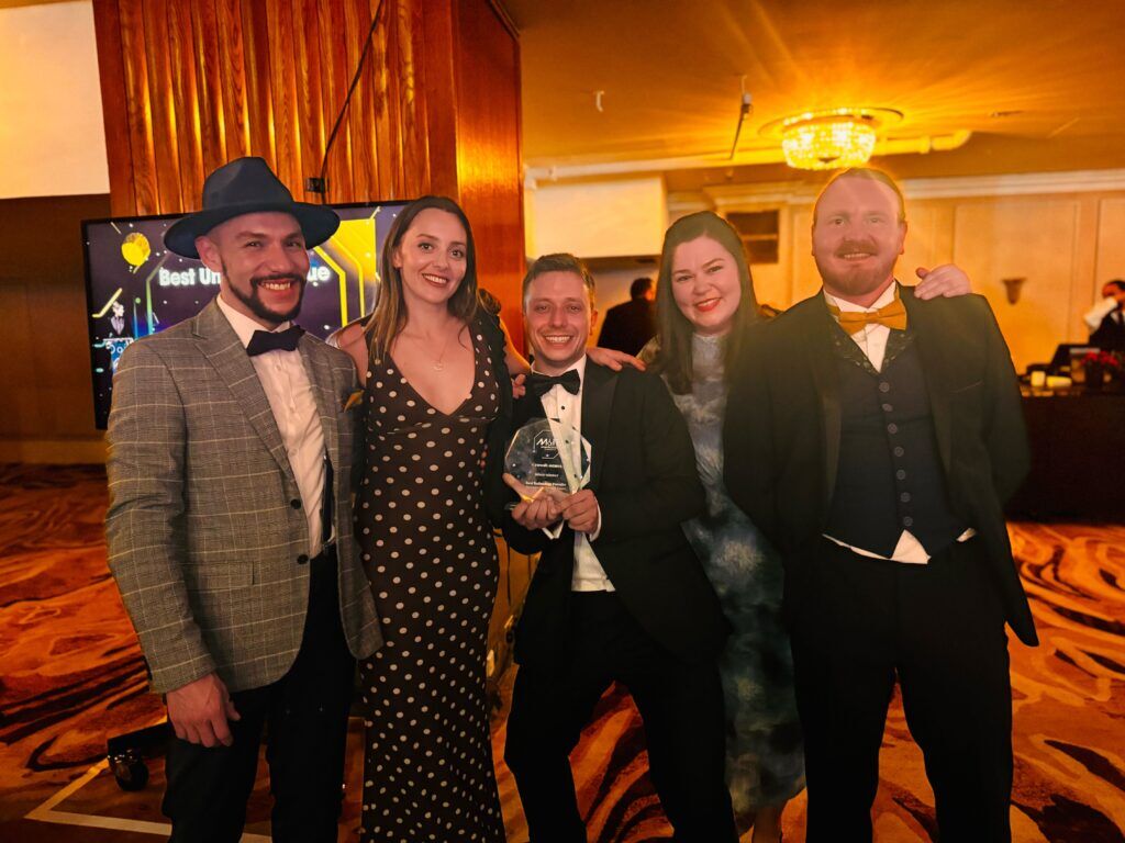 Five people in black tie at an awards dinner posing together with a glass trophy, a category screen lit up behind them