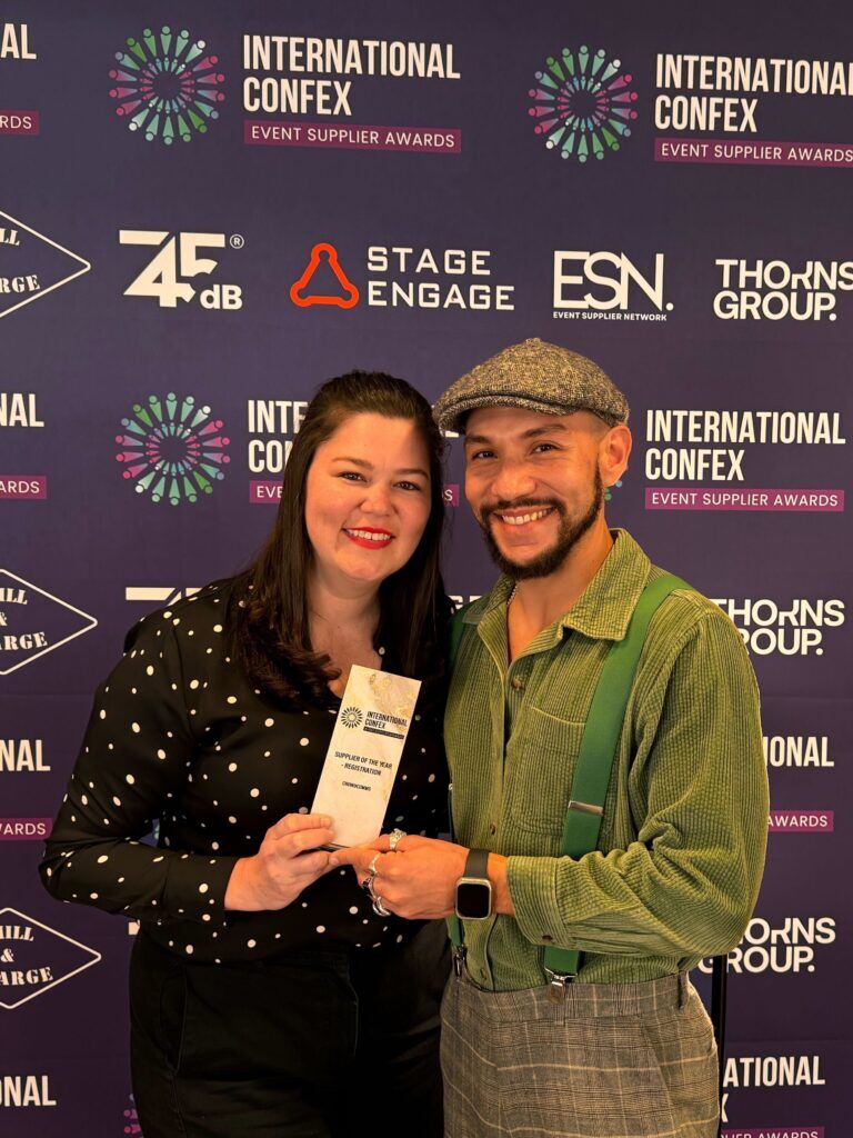 Two colleagues holding a "Supplier of the Year, Registration" trophy in front of an International Confex Event Supplier Awards backdrop