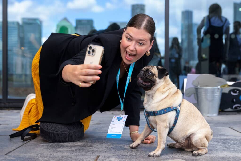 Woman in an Attendee lanyard crouching to take a phone selfie with a pug on a rooftop terrace, city skyline behind
