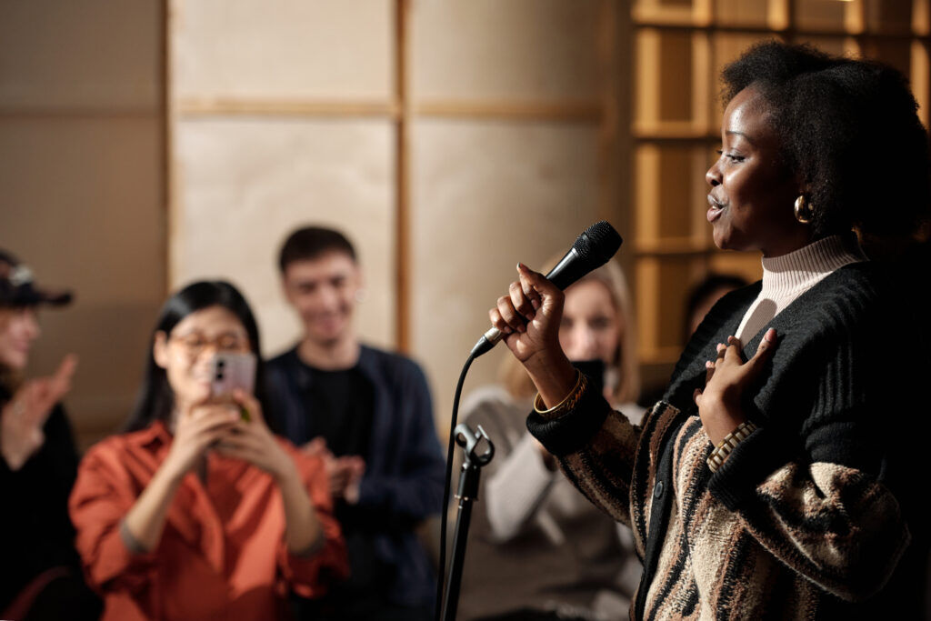 Woman speaking into a handheld microphone while a seated audience applauds and films her on a phone