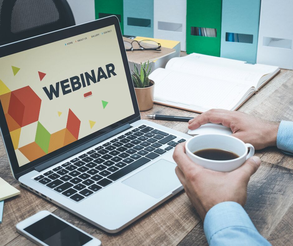 Man holding a coffee cup at a laptop showing a web page with the word WEBINAR in large type