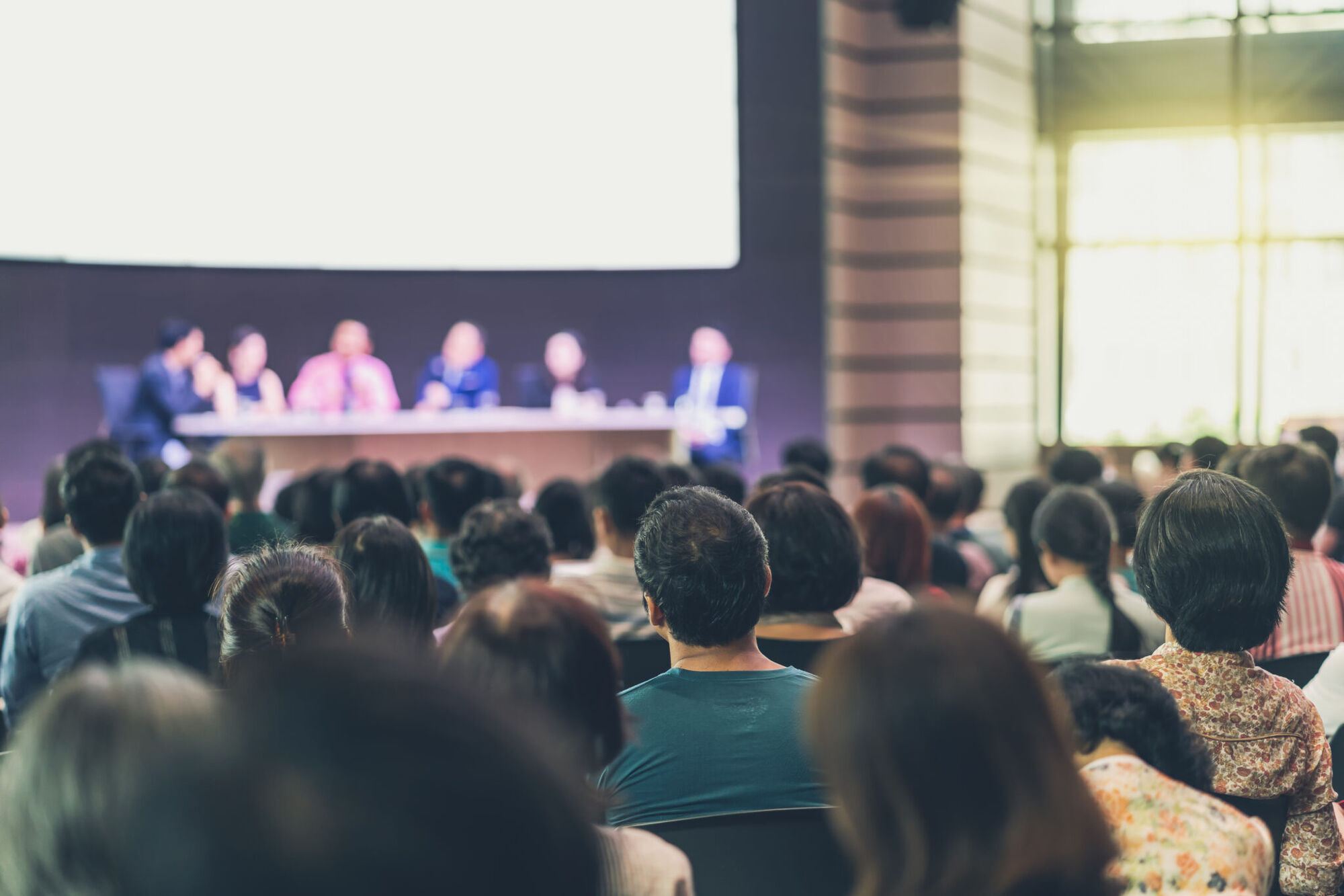 Packed conference audience seen from behind, facing a blurred panel of speakers at a table below a blank projection screen