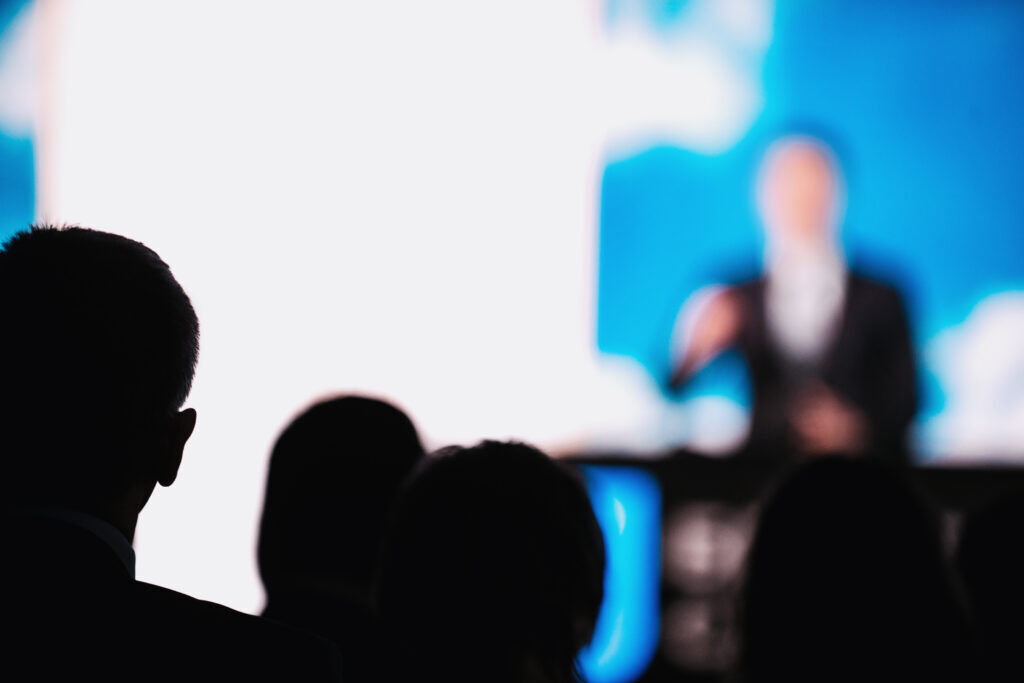 Silhouetted heads of an audience in front of a bright screen showing a blurred speaker at a lectern