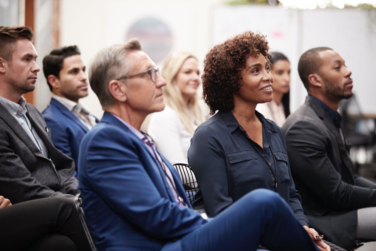 Row of smartly dressed people seated in an audience, listening and looking towards a speaker off camera