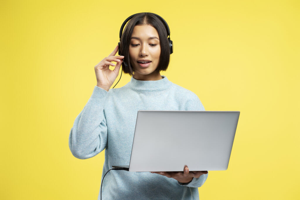 Woman in a pale blue jumper and headset holding an open laptop, against a plain yellow background
